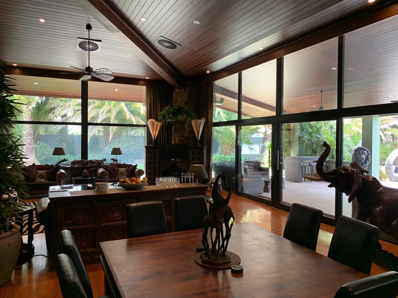 Jamala Lodge dining area, the decor is dark wood and safari inspired including a wooden elephant statue, the floor-to-ceiling windows look and onto a covered decking with outdoor furniture