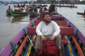 Tony sitting in the middle of a brightly multicoloured open boat with his red backpack between his knees and a sun hat on, the driver sports at the back of the boat behind Tony, in the background by many different coloured boats on the river, some with fresh produce. There is also some houses on the riverbank and a footbridge across the river.