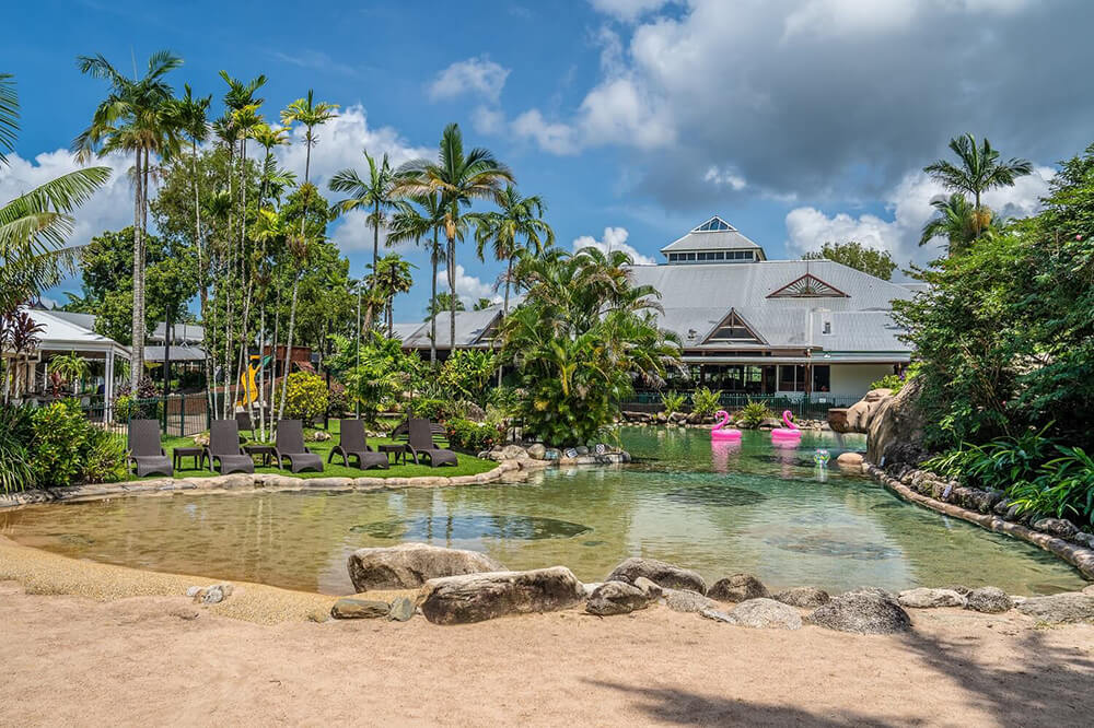 A lagoon type swimming pool surrounded by sand and rocks and trees, on the left are some sun lounges and in the background is a large building with grey roof, in the swimming pool our two inflatable pink flamingos