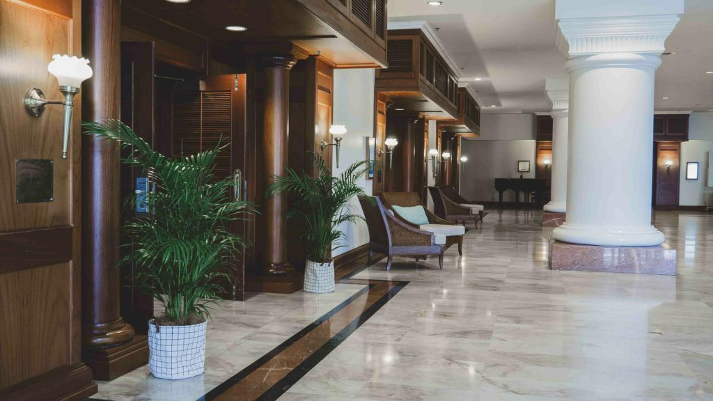 Colonial style hotel foyer with huge white columns in the middle of a marble floor to the left is dark wood panelling and venetian doors opening into a room. There are green plants and lights along the wall, there are also some chairs and at the very far end of the piano