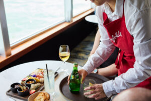 Waitress serves food and drinks to passenger at an indoor table overlooking the water 300x200